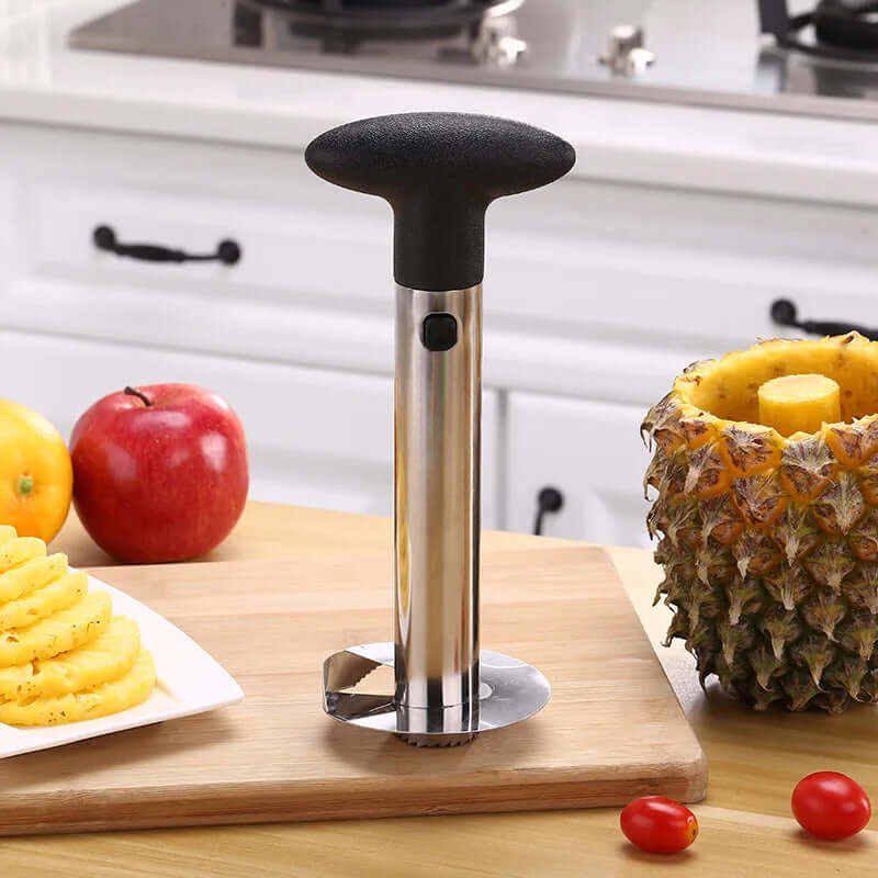 Stainless steel pineapple peeler cutter corer on a kitchen counter with sliced pineapples and apples in the background.