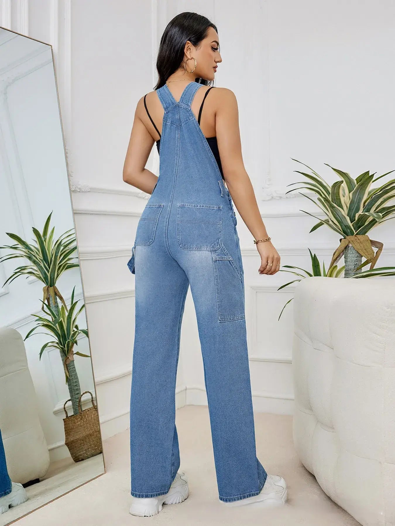 Back view of a woman in a denim bib jumpsuit, showcased in a stylish indoor setting.